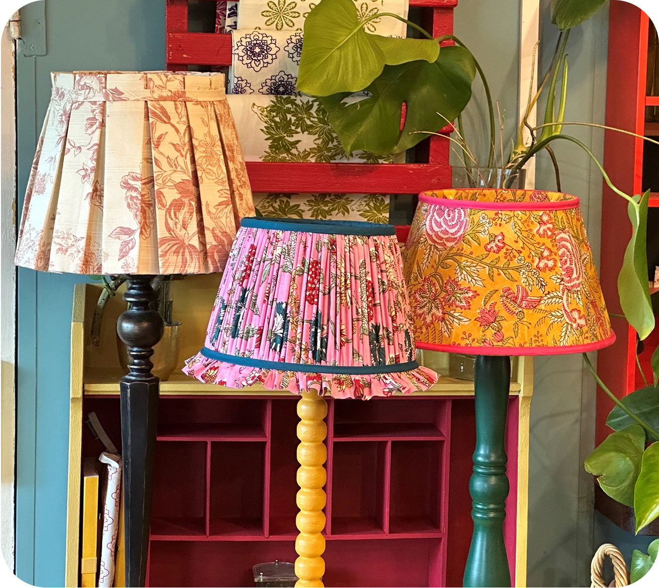 Three table lamps with colorful, patterned shades stand in a row against a brightly decorated background. The lampshades feature floral and pleated designs.